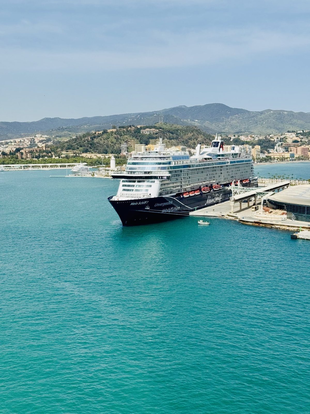 Ein großes Kreuzfahrtschiff liegt an einem Pier in einem sonnigen Hafen mit türkisfarbenem Wasser und grünen Hügeln im Hintergrund