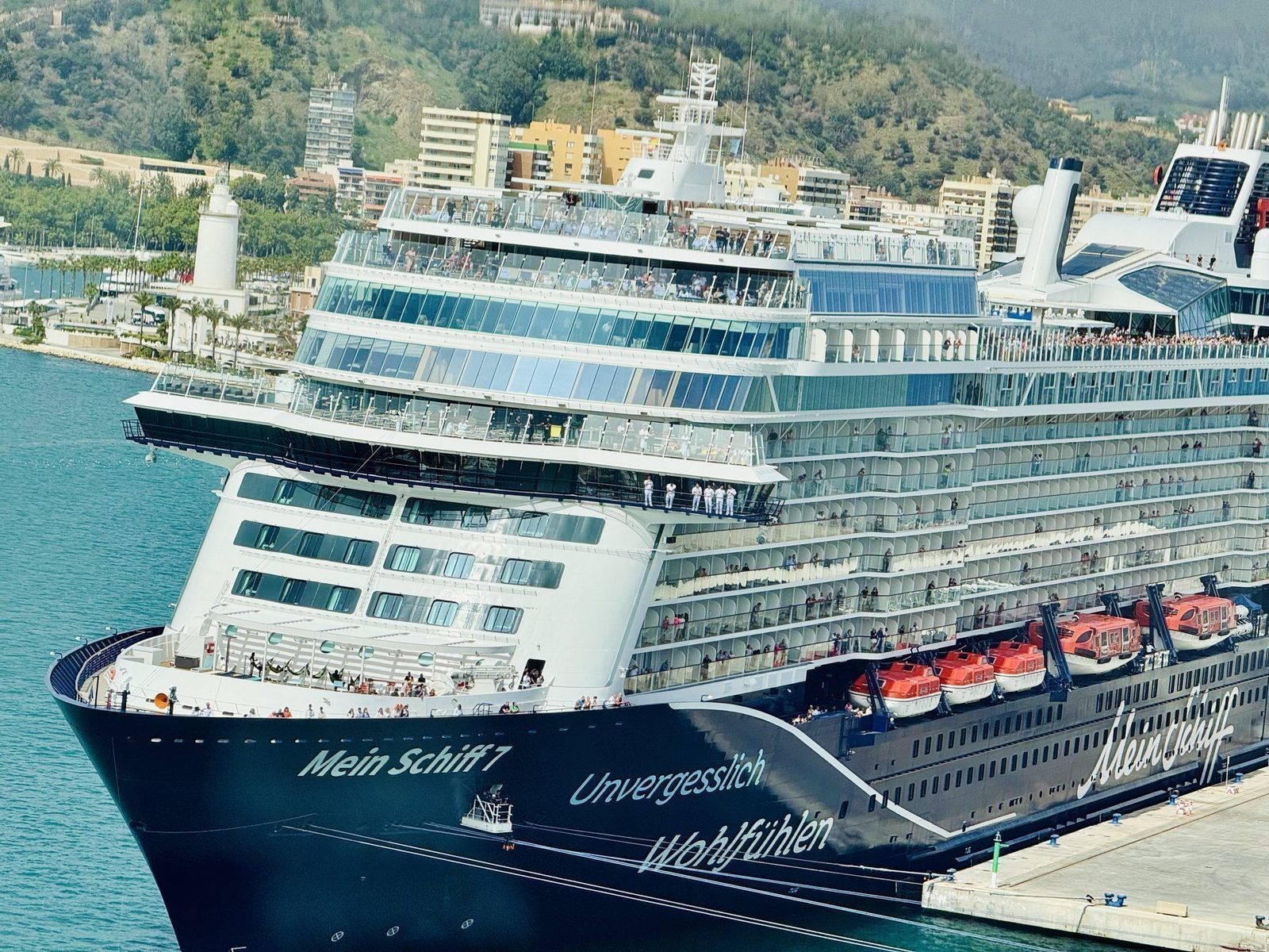Ein großes Kreuzfahrtschiff mit dem Namen 'Mein Schiff 7' ist im Hafen zu sehen, auf dessen Deck Menschen sich versammelt haben